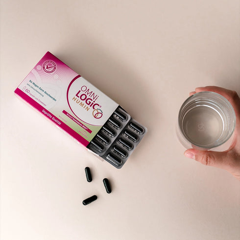 Eine offene Packung OMNi-LOGiC HUMIN mit losen Kapseln und einem Glas Wasser